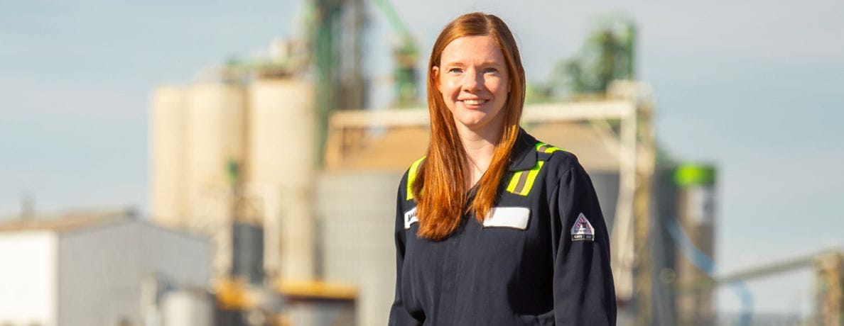 Woman in safety gear standing outdoors at an industrial site, smiling at the camera with equipment in the background.