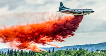 Aircraft dropping red fire retardant over a forest during wildfire suppression efforts.