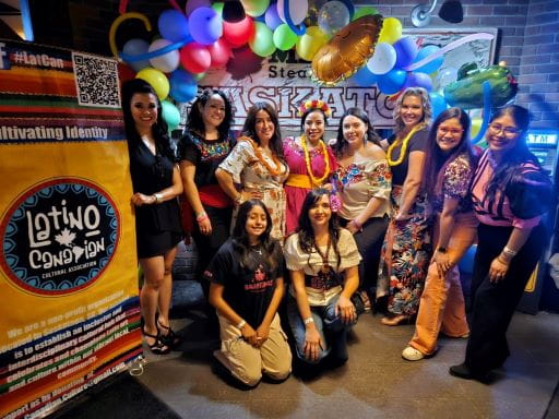 A group of women standing beside a banner with colourful balloons in the background for Latino Canadian Cultural Association. The group was one of a number that took part in the artsvest Saskatchewan program. Program applications are now open.