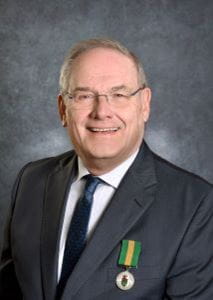 William Brooks, wearing a dark suit and glasses with the Saskatchewan Volunteer Medal pinned to his jacket. 
