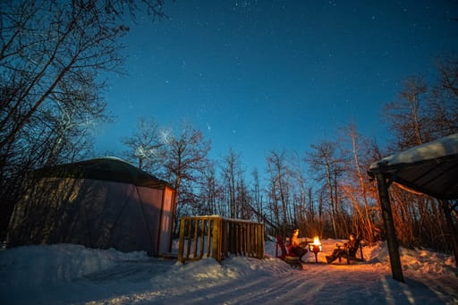 Two people sit around a campfire in winter with a yurt to the left