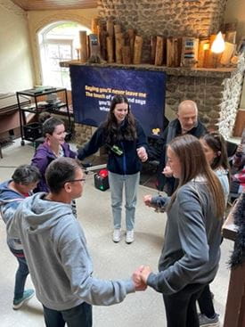 A group of campers and staff stand in a circle indoors, holding hands. A large screen in the background displays karaoke lyrics, and the room has stone walls and warm lighting.