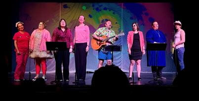 A group of performers in colorful outfits singing on stage, with one person playing guitar, under vibrant stage lighting.
