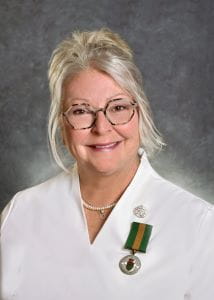 Brenda Corman, wearing glasses and a white blouse, the Saskatchewan Volunteer Medal is pinned to her blouse.