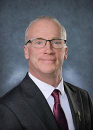 Mr. Brad Crassweller, MLA for White City-Qu’Appelle wearing a dark blue suit, white shirt and red tie, along with glasses. 