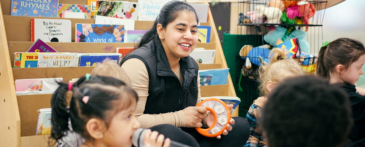 An early childhood educator is sitting on the floor surrounded by young children. She is playing the tambourine, a child in the forefront of the photo is holding a microphone.