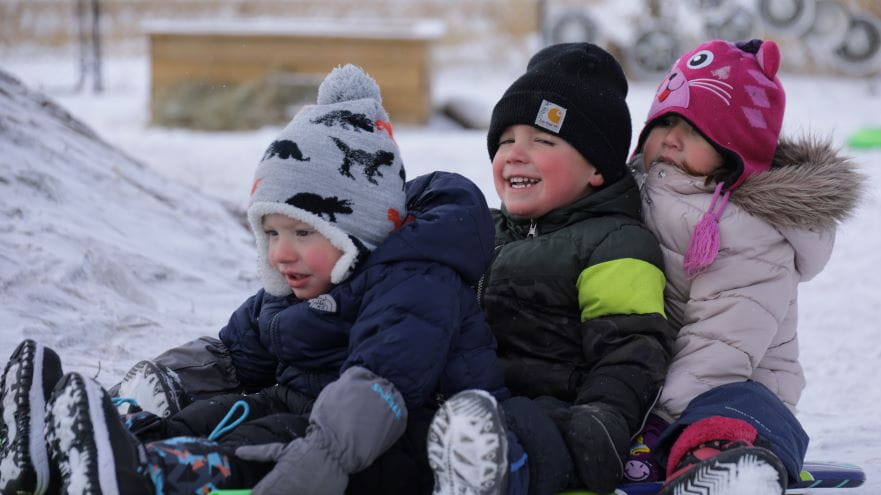 Three children are sitting together outside on a toboggan. The children are wearing winter clothing of hats, jackets and winter boots. They are cheerful as they play outside.