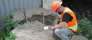 Engineer in an orange vest and hardhat reviewing notes while kneeling at a job site.