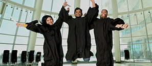 Three students, two female and one male, wearing black grad gowns, holding rolled up diplomas and jumping in front of some windows.