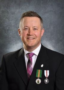 John Grant, wearing a black suit jacket and purple tie. The Saskatchewan Volunteer Medal is pinned next to another medal on his suit jacket. 
