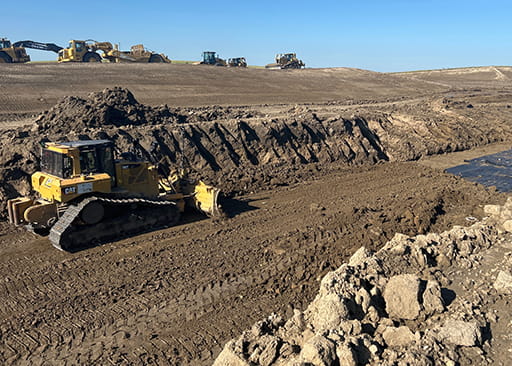 A yellow bulldozer moves brown dirt on a construction site with other heavy equipment in the background and a blue sky.