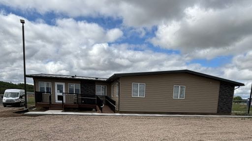 A single-story modular building with beige siding and dark trim, featuring a front porch with steps and a wheelchair-accessible ramp. The building sits on a gravel lot under a partly cloudy sky.