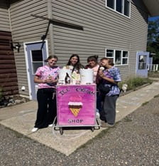 Four staff stand behind a brightly painted pink sign that reads “Ice Cream Shop,” holding ice cream cones. The group is smiling and standing outside on a sunny day near a building with chalk drawings on the sidewalk.