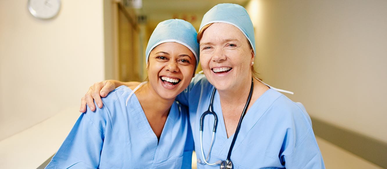 Two female health care workers wearing blue scrubs and head coverings, smiling