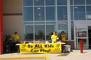Volunteers in yellow shirts running a fundraising booth with a banner that reads 'So ALL Kids Can Play!' outside a store.