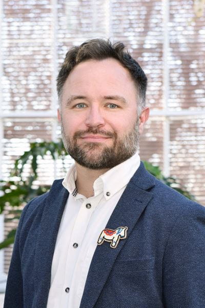 Person wearing a white button-up shirt under a textured navy blazer, accessorized with a decorative embroidered horse-shaped pin on the left lapel. The background shows softly lit windows with a blurred view of greenery.