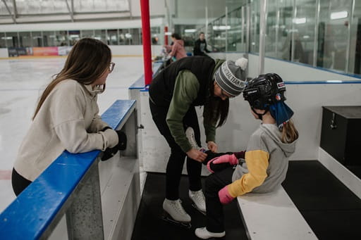 Young girl on a bench having her skates tied at an indoor arena. The girl is wearing a black helmet and they are on the bench at the skating rink. The Community Rink Affordability Grant Program application period is now open.