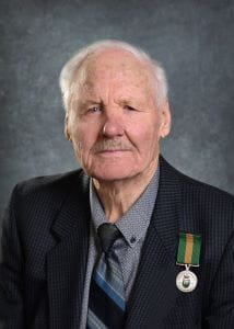Oswald Lutz, wearing a black suit jacket and grey, black and blue tie. The Saskatchewan Volunteer Medal is pinned to his suit jacket. 