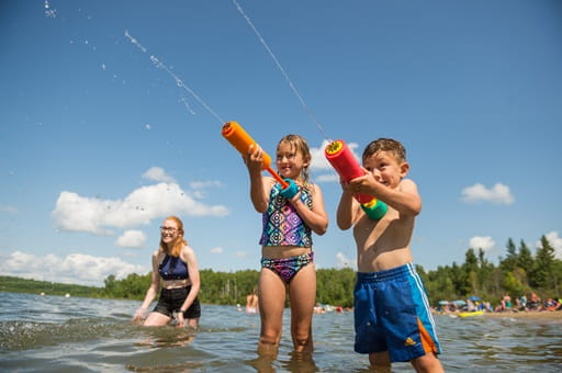 Three children are playing in shallow water at a beach, using colorful water guns to spray water. In the background, there are trees, people enjoying the beach, and a clear blue sky with some clouds.