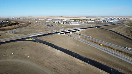 A highway overpass at Moose Jaw with one new bridge with new black pavement over a road and an existing bridge with faded pavement over a road. A bright blue Saskatchewan sky is in the background.