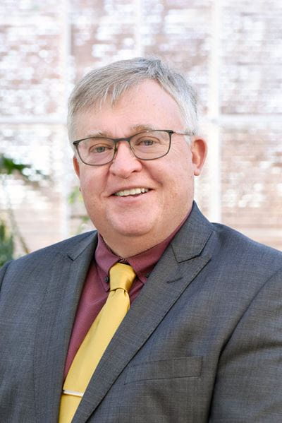 Person wearing a gray suit jacket over a maroon dress shirt, paired with a bright yellow necktie and a silver tie clip. The background shows a softly lit indoor setting with large windows and blurred greenery.