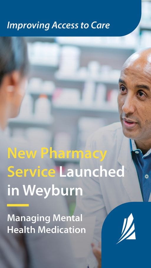 Male pharmacist in white lab coat leans forward from behind a pharmacy counter to discuss medication with a client. The client's face is not visible to the viewer.