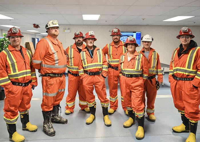 Ten NSEQC members, wearing orange coveralls with reflective stripes, hardhats with lights and heavy steel-toed boots, stand in a meeting room as they await a tour of Foran's McIlvenna Bay Mine while it's under construction. 