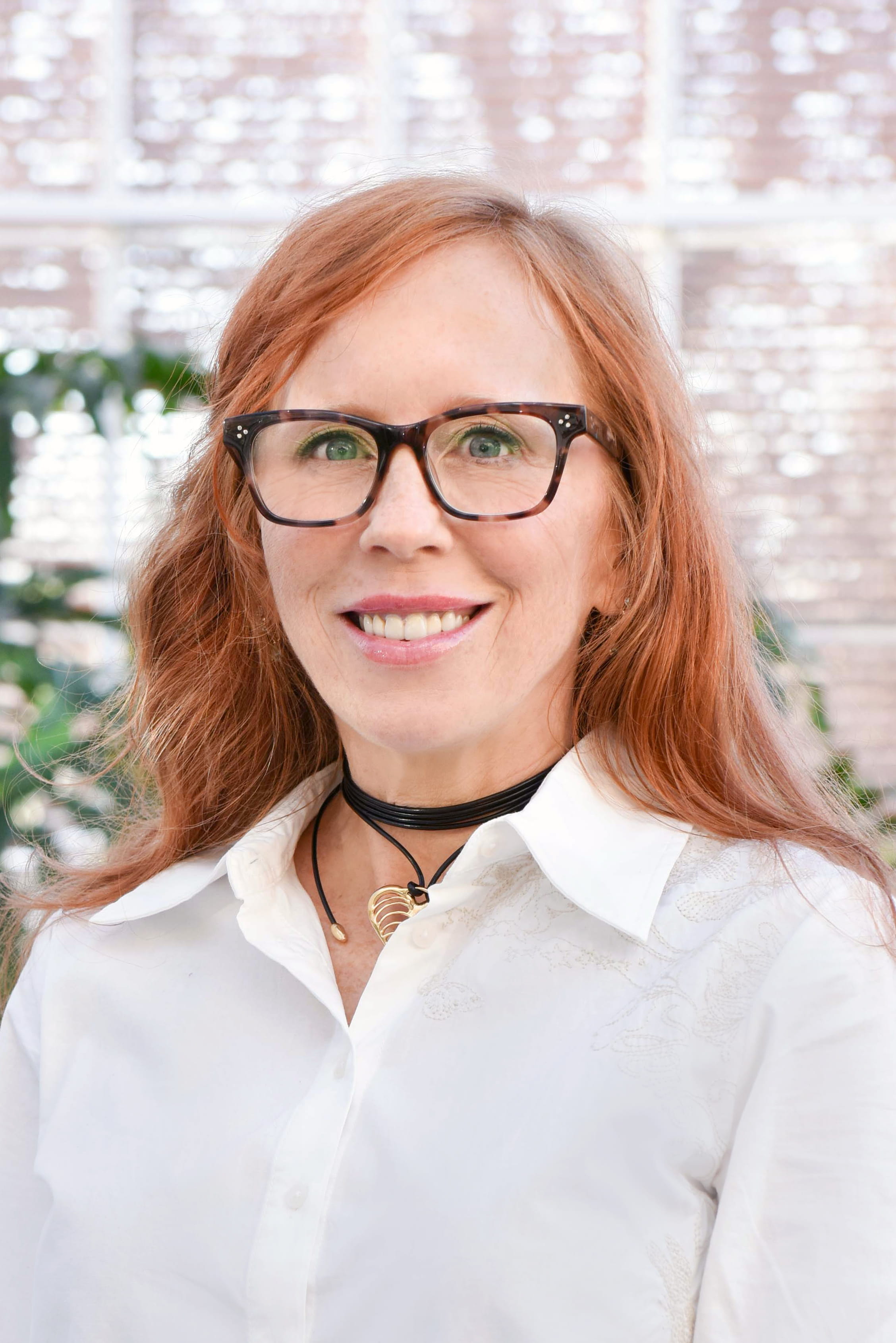 Person wearing a white collared shirt with subtle embroidered details, accessorized with layered black choker necklaces and a gold pendant. The background features softly lit windows and blurred greenery.