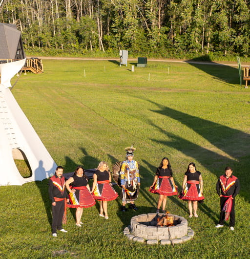 A group of people dressed in traditional Indigenous regalia stand in a semi-circle behind a firepit. A tipi is pictured behind them. Pêmiska Tourism is a finalist at the 2026 Indigenous Tourism Awards, which will be held in Edmonton on February 19, 2026.