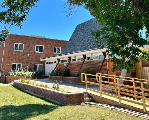Photo of Pine Lodge Addiction Recovery, located at 3535 8th Avenue in Regina. It depicts a brown brick building with a grey V-domed roof. A wooden ramp with railings leads up to the building. Connected to this building is a two-storey brick building with white-framed windows. The photo is taken in summer, depicting green grass and a blue sky. The sober living spaces are located at two homes separate from this main facility.