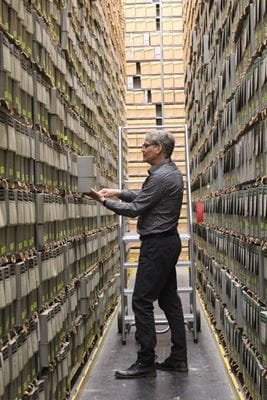 An older man wearing black pants and grey shirt stands in narrow corridor between two-metre high shelving units filled with files, books and papers at the Provincial Archives of Saskatchewan.