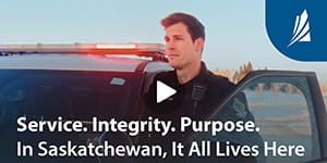 A police officer stands between a vehicle and its open door. A Saskatchewan logo is in the top right corner on a blue background. The text at the bottom reads Service. Integrity. Purpose. In Saskatchewan, It All Lives Here.