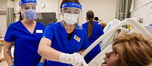 Two female nursing students wearing blue scrubs, face masks and plastic face coverings working in a nursing simualtion lab.