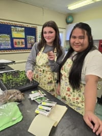 Two teenage girls donning colourful gardening aprons smile while standing behind a workbench in a science classroom. A tray of seedlings sits on the workbench beside several small bags of vegetable seeds. The girl further from the camera holds up a small seedling in her left hand, showing it off to the photographer.