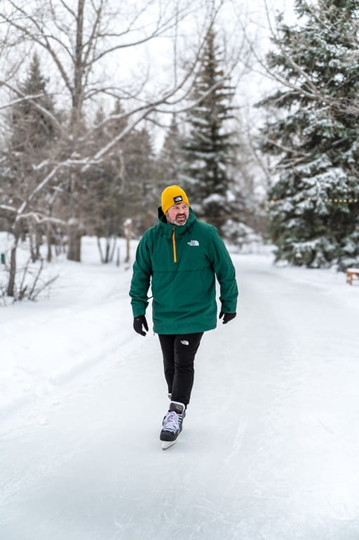 A man in a green jacket and yellow toque skates outside.