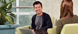 Two students facing each other, sitting in green chairs with a plant and a window behind them. Both students have a laptop in their laps. 