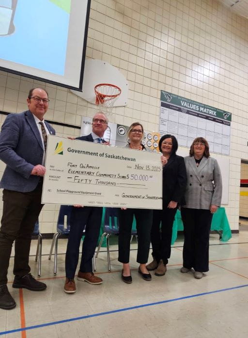 Five people inside a gymnasium holding a large white cheque for the School Playground Equipment Grant program for Fort Qu’Appelle Elementary Community School for $50,000. The big cheque has the green and yellow Government of Saskatchewan logo on it. There is an orange basketball hoop on the wall behind the group.