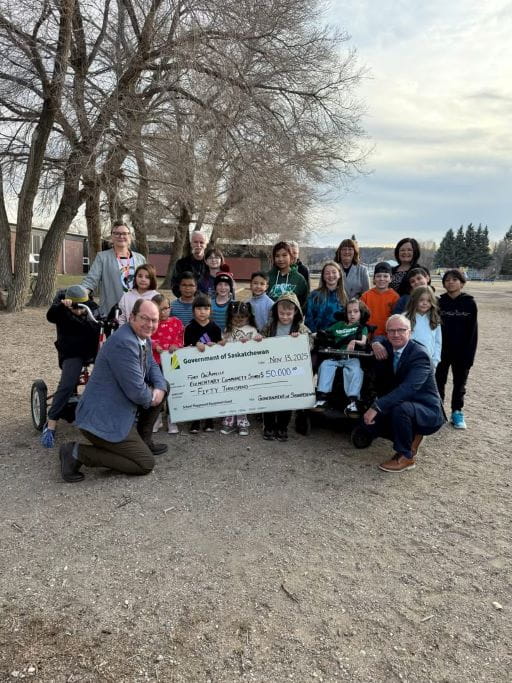 A group of elementary students and adults outside with a large white cheque for the School Playground Equipment Grant program for Fort Qu’Appelle Elementary Community School for $50,000. The big cheque has the green and yellow Government of Saskatchewan logo on it. There are trees in the background and the sky behind the group is blue.