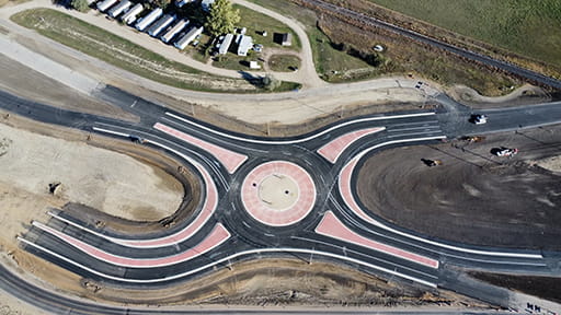 A traffic circle known as a roundabout at Weyburn under construction with new black pavement. A concrete border outlines a circle in the middle. Equipment is nearby.