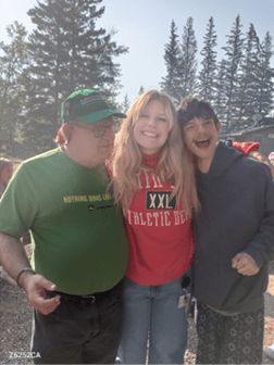 A staff member and two campers posing for a picture, smiling with trees in the background. The person in the middle has their arms around the two others.